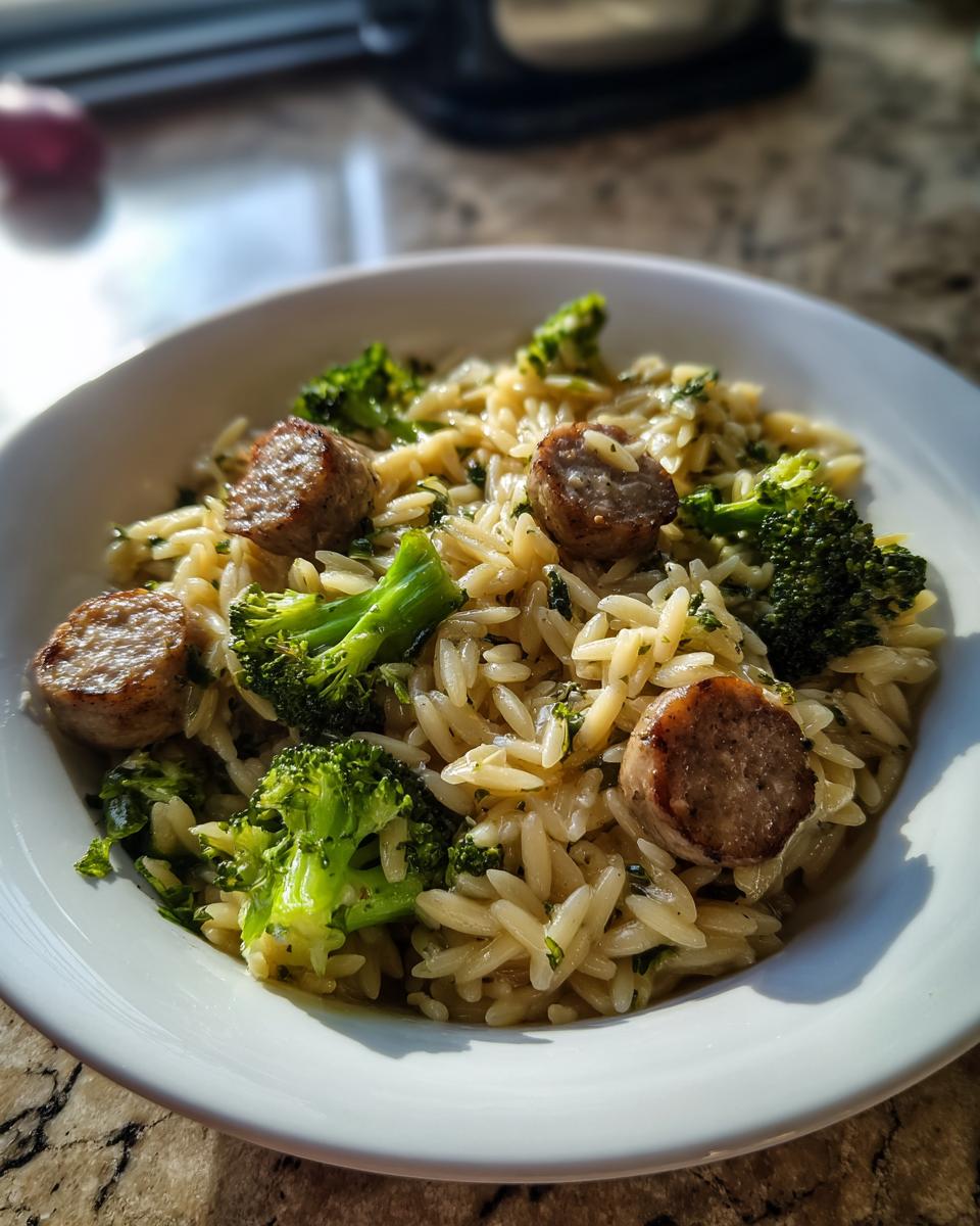 Close-up of chicken sausage and broccoli orzo in a white bowl, well-lit by natural light.