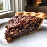 A close-up of a rich slice of chocolate bourbon pecan pie with a flaky crust, served on a white plate.