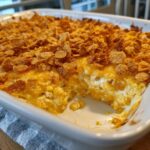 A scoop has been taken out of a baked cream cheese corn casserole topped with melted cheese and crunchy cornflakes.