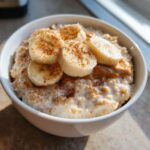 A close-up of creamy banana oatmeal topped with sliced bananas, a swirl of peanut butter, and a dusting of cinnamon.