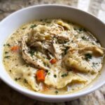 Close-up of a white bowl filled with creamy chicken and dumplings, topped with shredded chicken and parsley.