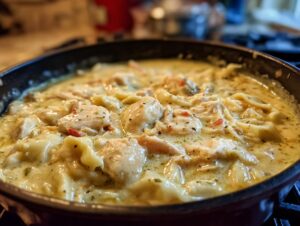Close-up of creamy chicken and dumplings simmering in a dark skillet, featuring tender chicken pieces and flat noodles.
