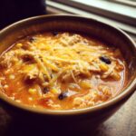 Close-up of a bowl of creamy chicken enchilada soup topped with shredded cheese, corn, and black beans.