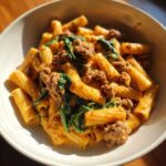 Close-up of creamy sausage pasta mixed with wilted spinach served in a light-colored bowl.