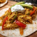 Close-up of a soft tortilla loaded with shredded crockpot chicken fajitas, bell peppers, avocado slices, and sour cream.