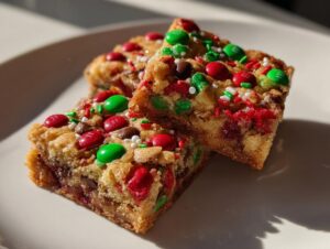 Close-up of two stacked, chewy christmas cookie bars topped with red and green M&Ms and holiday sprinkles.