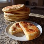 A stack of fluffy homemade pita bread with two halves cut open on a white plate in the foreground.