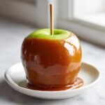 A glossy, homemade caramel apple with a wooden stick, resting on a small white saucer.
