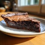 A rich, gooey slice of hot fudge pie sits on a light-colored plate, catching the sunlight.