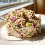 A mound of creamy healthy tuna salad mixed with diced green celery and purple red onion, served on a white plate.