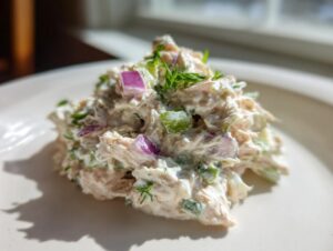 A mound of creamy healthy tuna salad mixed with diced red onion and celery, topped with fresh dill on a white plate.