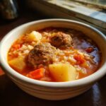 Close-up of a white bowl filled with rich, tomato-based albondigas soup featuring meatballs, rice, potatoes, and carrots.