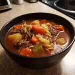 Close-up of a black bowl filled with rich vegetable beef soup, featuring chunks of beef, carrots, potatoes, and egg noodles.