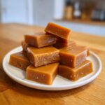 Close-up of several squares of rich, chewy homemade caramel stacked on a small white plate.