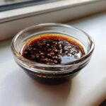 Close-up of rich, dark homemade teriyaki sauce topped with toasted sesame seeds in a small glass bowl.