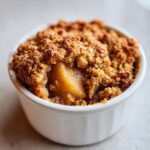 Close-up of a warm, individual apple crumble in a white ramekin, showing the golden brown topping and soft cooked apple filling.