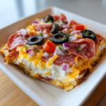 A close-up, square serving of loaded hoagie dip topped with pepperoni, shredded cheddar, black olives, and red onion.