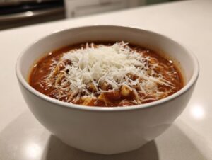 A close-up of a white bowl filled with rich, tomato-based minestrone soup topped generously with shredded Parmesan cheese.