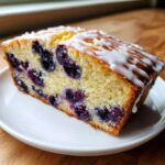 A close-up of a moist slice of blueberry bread, studded with dark berries and topped with a white sugar glaze.