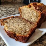 Close-up of a moist slice of sourdough banana bread next to the main loaf on a white plate.