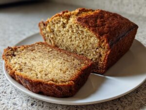 A close-up of a moist sourdough banana bread loaf, with one thick slice cut and resting beside it on a white plate.