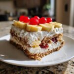 A delicious square slice of no-bake banana split cake featuring layers of graham cracker crust, cream, bananas, and topped with cherries.