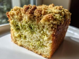 A close-up, sunlit slice of pistachio coffee cake showing a crumbly streusel topping and green pistachio bits throughout the moist crumb.