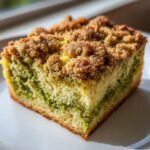 Close-up of a moist slice of pistachio coffee cake featuring a green pistachio swirl and a golden brown streusel topping.