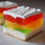 Close-up of a square slice of rainbow jello poke cake showing layers of white cake, green, yellow, and red gelatin, topped with white whipped cream.