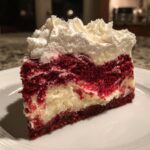 A thick slice of red velvet cheesecake with marbled red cake and cream cheese layers, topped with whipped cream.