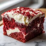 Close-up of a square slice of moist red velvet poke cake topped with thick cream cheese frosting and red crumbs.