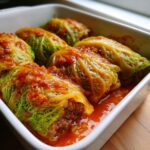 Close-up of homemade cabbage rolls stuffed with meat, simmering in a rich tomato sauce in a white baking dish.
