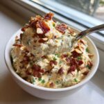 A spoonful of creamy Neiman Marcus dip topped with bacon bits and almonds being lifted from a white bowl.