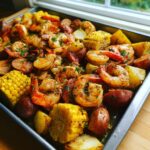 Close-up of a sheet pan filled with seasoned shrimp boil ingredients including shrimp, corn on the cob pieces, red potatoes, and sausage.