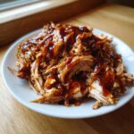 Close-up of tender, shredded crockpot teriyaki chicken generously drizzled with glossy teriyaki sauce on a white plate.