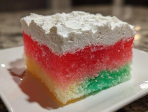 Close-up of a layered slice of jello poke cake showing yellow, green, and red gelatin absorbed into the cake, topped with white whipped cream.