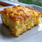 A close-up of a moist slice of cornbread casserole with visible corn kernels and a golden, slightly caramelized top.