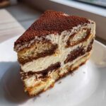 A perfect slice of tiramisu cheesecake showing layers of cream, ladyfingers, and a heavy dusting of cocoa powder on top.