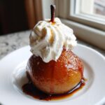 A beautifully glazed, whole poached pear topped with a swirl of whipped cream and syrup on a white plate.