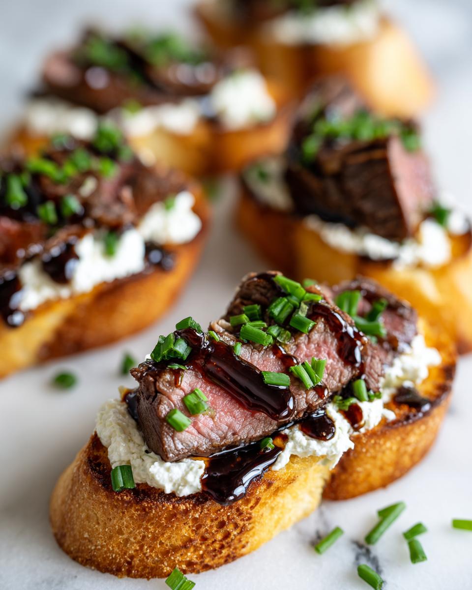 Close-up of a steak crostini topped with creamy cheese, medium-rare steak, dark glaze, and fresh chives.