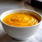 Close-up of a steaming bowl of vibrant orange cheese soup, garnished with black pepper.