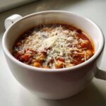 Close-up of a steaming white mug filled with rich minestrone soup topped generously with shredded Parmesan cheese.