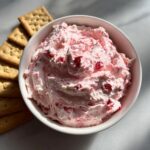 A white bowl filled with fluffy pink strawberry cheesecake dip, served alongside square crackers.