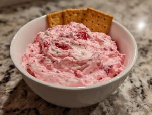 A white bowl filled with creamy pink strawberry cheesecake dip, served with graham crackers sticking out.
