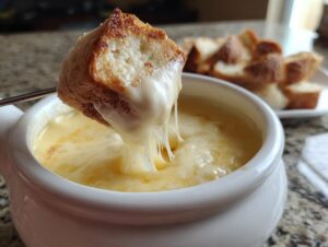 A piece of crusty bread being dipped into a white bowl of hot, stretchy cheese fondue.