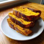 Four thick slices of vibrant orange sweet potato toast, slightly charred, stacked on a light gray plate.