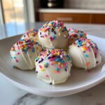 A pile of white chocolate coated sugar cookie truffles topped with colorful rainbow sprinkles on a white plate.