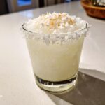 A close-up of a frosty white Christmas margarita in a rocks glass, rimmed with coconut and topped with toasted coconut flakes.