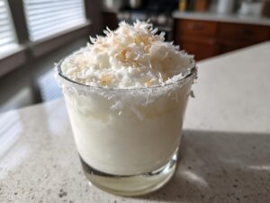 A creamy, white Christmas margarita in a short glass, rimmed with coconut and topped with toasted coconut flakes.