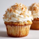 A close-up of a moist coconut cupcake topped with white frosting and toasted coconut flakes.